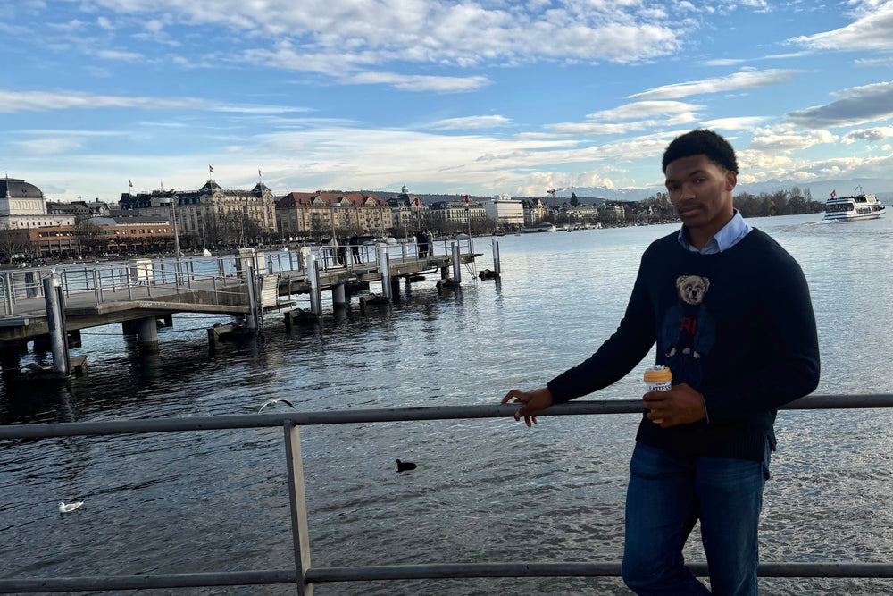 A man wearing a navy blue sweater, blue jeans, and brown shoes stands on a waterfront balcony holding a coffee cup, with a pier, water, and a city skyline under a cloudy blue sky in the background.