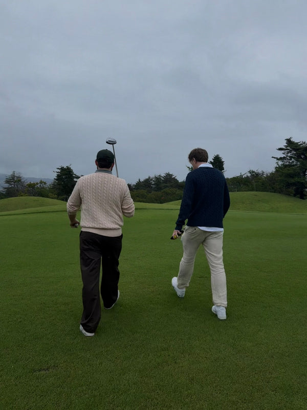 Two men seen from behind walking on a golf course under an overcast sky, one wearing a beige knit sweater and dark trousers, and the other wearing a navy sweater, light trousers, and white shoes.