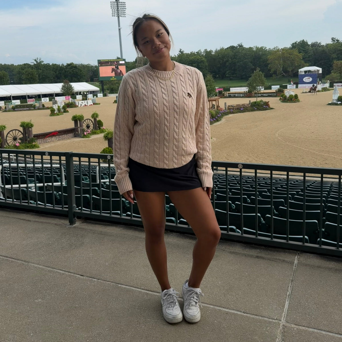 A young woman stands on an outdoor concrete walkway in front of a black metal railing, wearing a light beige cable-knit sweater, a black mini skirt, and white sneakers. The background shows a horse riding arena under a cloudy sky.