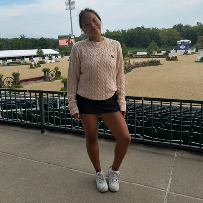 A young woman stands on an outdoor concrete walkway in front of a black metal railing, wearing a light beige cable-knit sweater, a black mini skirt, and white sneakers. The background shows a horse riding arena under a cloudy sky.