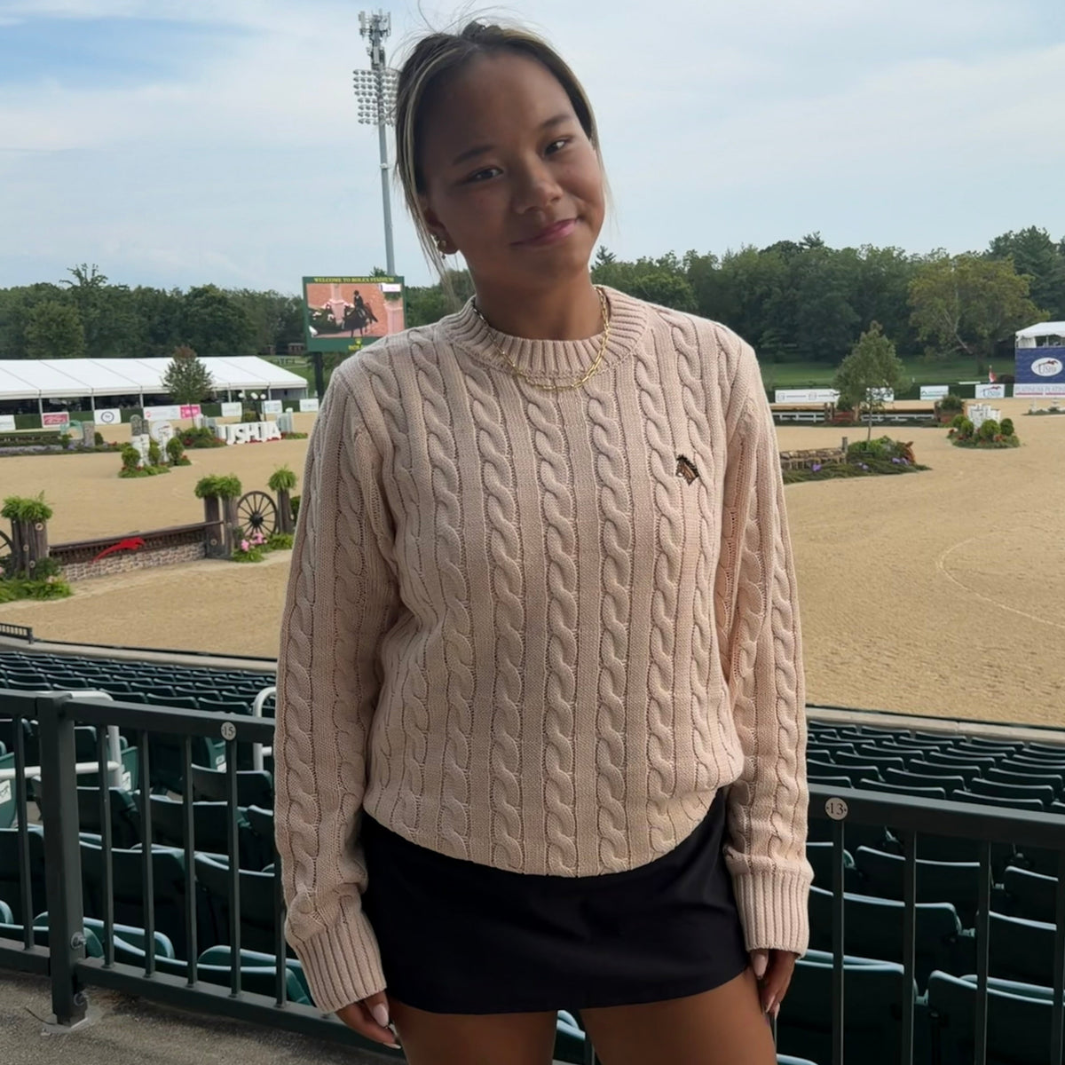 A young woman stands outdoors at an equestrian arena, wearing a light beige cable-knit sweater with a small embroidered logo on the chest and black athletic shorts.