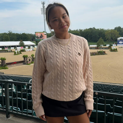 A young woman stands outdoors at an equestrian arena, wearing a light beige cable-knit sweater with a small embroidered logo on the chest and black athletic shorts.