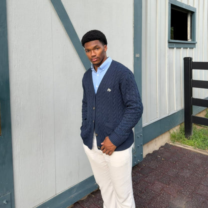 A man wearing a navy blue cable-knit cardigan over a light blue collared shirt and white pants, standing in front of a white barn with dark teal trim.